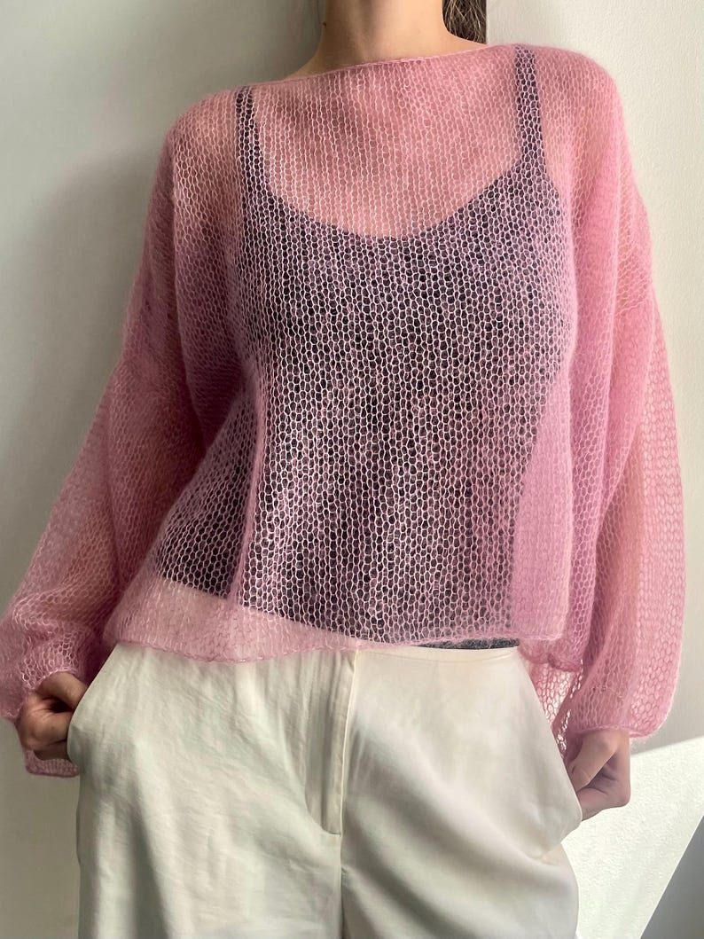 Puede incluir: Un su&eacute;ter de punto rosa claro y transparente con cuello ancho y mangas largas. El su&eacute;ter se usa sobre una camiseta negra y se combina con pantalones blancos. El tejido tiene un tejido delicado y abierto.