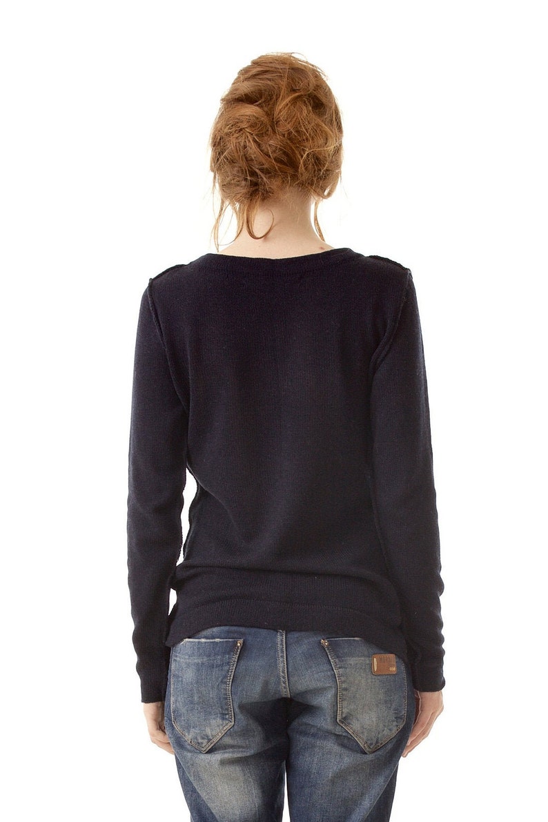 May include: A long-sleeved, dark navy blue knit sweater with a round neckline, paired with blue denim jeans featuring a brown leather patch on the back pocket. The sweater has a simple, solid-colored design with ribbed cuffs and a ribbed hemline.