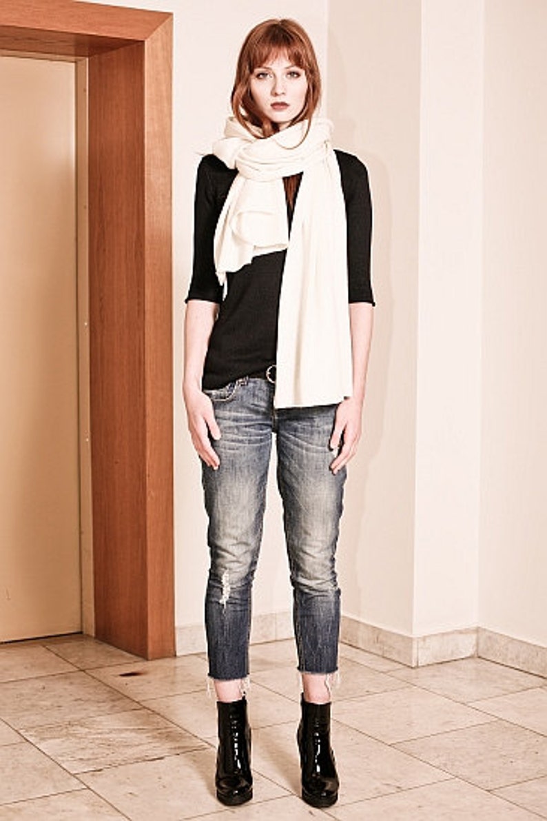 May include: A woman wearing a white scarf, a black long-sleeved shirt, blue jeans, and black ankle boots.
