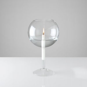 Puede incluir: Un portavelas de cristal transparente con una parte superior esférica y un tallo delgado. Una vela blanca está encendida dentro del cristal, con la llama visible. El portavelas está sobre una superficie blanca sobre un fondo gris.