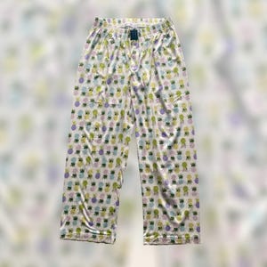 May include: White pajama pants with an all-over pattern of colorful pineapples in shades of yellow, green, and purple. The pants have an elastic waistband and a drawstring. The fabric appears to be lightweight and silky.