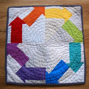 May include: A square, quilted wall hanging with a repeating arrow design. The arrows are in a rainbow of colors: red, orange, yellow, green, teal, blue, and purple. The background is white with concentric circle quilting.