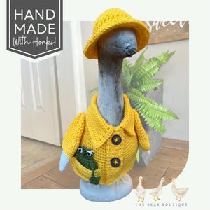May include: A whimsical goose statue wearing a handmade yellow crochet rain jacket and hat. The jacket has two brown buttons and a pocket with a green frog. The text "HAND MADE With Honks!" is in the upper left corner.