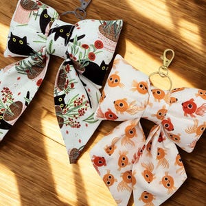 May include: Two fabric bow keychains. One features a white background with black cats and potted plants. The other has a white background with orange goldfish. Both have metal clasp attachments and are on a wooden surface.
