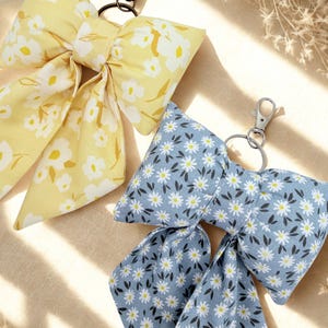 May include: Two floral bow keychains. One is yellow with white flowers, the other is blue with white daisies. Both have metal clasps for attaching to keys or bags. The bows are made of fabric.