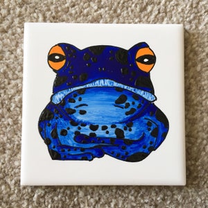 Könnte beinhalten: Handbemalte Keramikfliese mit einem blauen Frosch mit schwarzen Flecken und orangefarbenen Augen. Der Frosch sitzt auf einem weißen Hintergrund.