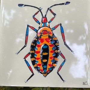 Könnte beinhalten: Quadratische Keramikfliese mit einer farbenfrohen Insektenillustration. Das Insekt hat einen schwarzen Körper mit orangefarbenen, blauen und roten Akzenten. Die Fliese hat einen hellbeigen Hintergrund und ist wahrscheinlich ein Dekorationsartikel.
