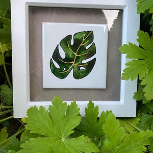 Pode incluir: Uma obra de arte emoldurada com um design de folha de monstera verde com detalhes dourados em um azulejo quadrado branco. A obra de arte está emoldurada em uma moldura de madeira branca e um passe-partout cinza, cercada por folhagem verde.