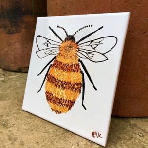 May include: A hand-painted ceramic tile depicting a bumble bee with black and yellow stripes and white wings. The bee is painted in a realistic style and the tile is signed "Elk" in the bottom right corner.