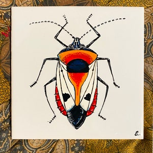 Op de afbeelding: Vierkante keramische tegel met een gestileerde insectenillustratie. Het insect heeft een zwarte kop, poten en antennes, met een fel oranje en rood lichaam. De achtergrond is wit en de tegel is rechtsonder gesigneerd.