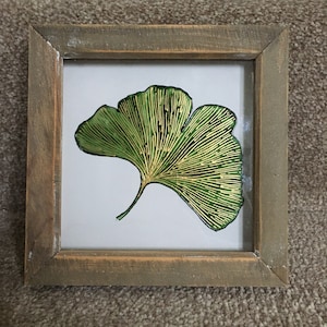 May include: A framed print of a green and gold ginkgo leaf. The leaf is detailed with gold veins and is set against a white background.