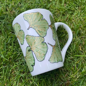 May include: White ceramic mug featuring a pattern of green and gold ginkgo leaves. The leaves are intricately detailed and outlined in black. The mug has a curved handle and is set on a bed of green grass.
