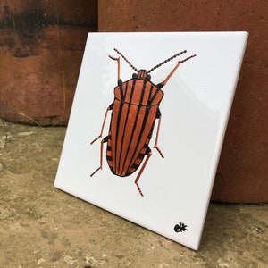 Op de afbeelding: Een handgeschilderde keramische tegel met een gedetailleerde illustratie van een kever met zwarte en oranje strepen. De kever is afgebeeld in een realistische stijl met ingewikkelde details.