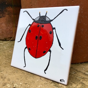 Può includere: Una piastrella di ceramica dipinta a mano con una coccinella rossa con macchie nere e gambe nere. La piastrella ha uno sfondo bianco ed è firmata "ell" nell'angolo in basso a destra.