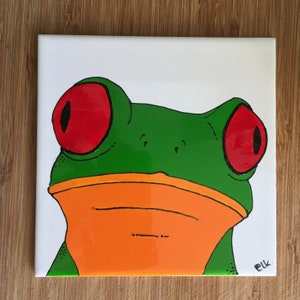 Könnte beinhalten: Nahaufnahme einer handbemalten Keramikfliese mit einem grünen Laubfrosch mit großen roten Augen und orangefarbenem Bauch. Der Frosch schaut direkt auf den Betrachter. Die Fliese ist mit "Elk." signiert.