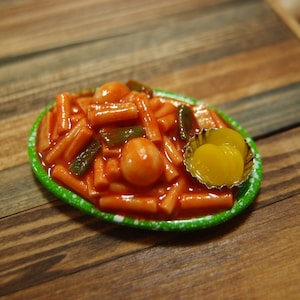 Puede incluir: Un plato en miniatura de tteokbokki coreano, con pasteles de arroz cilíndricos en una salsa roja vibrante, pimientos verdes y huevos duros. Un pequeño plato de acompañamiento de rábano encurtido amarillo se encuentra junto al plato principal. El plato tiene un borde verde con motas blancas.