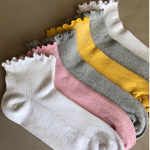 May include: A collection of ruffled-top socks in various colors, including white, pink, gray, and yellow. The socks are arranged in a diagonal line, showcasing their ribbed texture and delicate trim. The socks are made of a soft material.