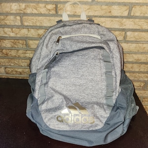 May include: A heather gray backpack featuring a large main compartment, a smaller front zippered pocket, and two side mesh pockets. The front displays a metallic gold Adidas logo and text. It includes a top carry handle and adjustable straps.
