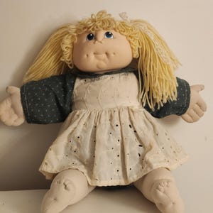 May include: A soft-sculpture doll with yarn blonde hair styled in pigtails, blue eyes, and dimpled cheeks. The doll wears a dark green long-sleeved top with a white floral pattern and a cream-coloured eyelet lace pinafore dress over the top.