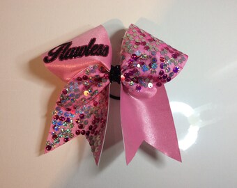 May include: Pink cheer bow with a glittery pink and silver sequin design. The bow has a black elastic band and the word "Flawless" is embroidered on one side in black.