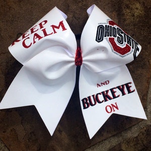 May include: White cheerleading bow with red glitter accents. The bow features the text "KEEP CALM" and "OHIO STATE AND BUCKEYE ON". The bow is a fan accessory.