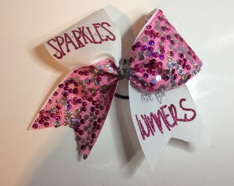 May include: A pink and white cheer bow with silver glitter. One side of the bow has pink sequins and the text "Sparkles are for winners" in pink glitter. The other side of the bow is white with the text "Sparkles" in pink glitter.