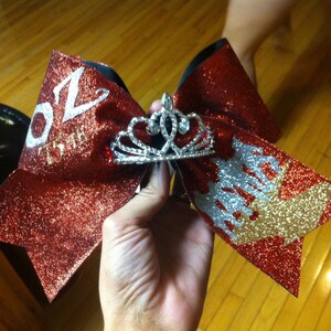 May include: A red glitter cheerleading bow with a silver tiara in the center. The bow has white text and a silver and gold design on the ends. The bow is held in a hand.