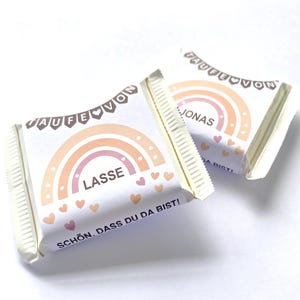 May include: Two wrapped candy bars with a rainbow design in shades of peach and orange. The bars have text that reads "LASSE" and "JONAS" with the phrase "SCH&Ouml;N, DASS DU DA BIST!" printed on the wrapper. The wrappers are white with a decorative banner.