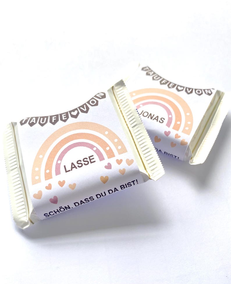 May include: Two wrapped candy bars with a rainbow design in shades of peach and orange. The bars have text that reads "LASSE" and "JONAS" with the phrase "SCH&Ouml;N, DASS DU DA BIST!" printed on the wrapper. The wrappers are white with a decorative banner.