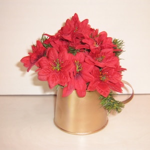 May include: A gold metal watering can with a red poinsettia flower arrangement. The flowers are artificial and have green leaves.