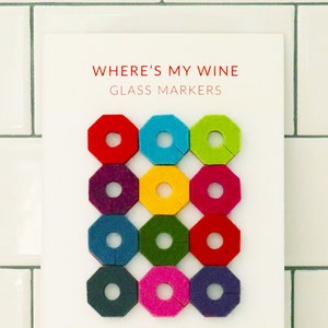 May include: A set of 10 colorful felt wine glass markers in various shades of red, green, blue, yellow, and purple. The markers are shaped like octagons with a hole in the center. The text "WHERE'S MY WINE GLASS MARKERS" is printed in red at the top of the card. The text "MINIMIZING WINE GLASS CONFUSION ONE VIBRANT COLOR AT A TIME" is printed in black below the markers. The text "BESPOKE PROVISIONS" is printed in red at the bottom of the card.