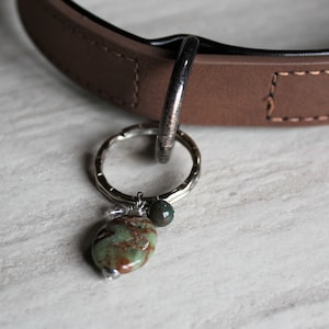 Puede incluir: Un collar de perro de cuero marrón con un anillo de plata y un dije con una piedra verde y marrón.