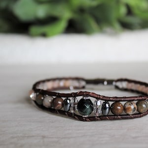 Könnte beinhalten: Ein braunes Lederarmband mit verschiedenen Edelsteinperlen, darunter grüne, braune und klare Perlen. Das Armband hat einen silbernen Verschluss.