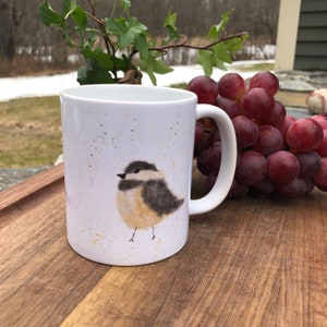 May include: White ceramic mug featuring a watercolor illustration of a small bird with black, gray, and tan feathers. The mug has a curved handle and is speckled with gold and brown flecks. The mug is on a wooden surface.