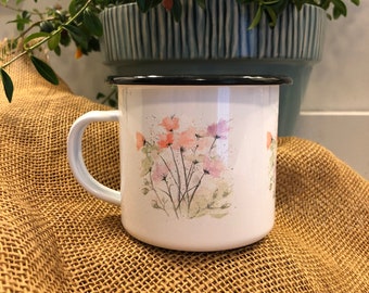 May include: White enamel mug with a black rim and a watercolor floral design featuring pink, orange, and green flowers. The mug is sitting on a brown burlap surface.