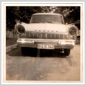 Puede incluir: Fotografía en blanco y negro de un coche Triumph antiguo, vista frontal. El coche tiene una parrilla cromada, faros y una matrícula que dice "B-LS 342". El coche está estacionado sobre una superficie pavimentada.