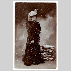 Postal vintage con foto real: Mujer, abrigo, sombrero, invierno, moda, glamour, nieve cayendo. Tarjeta de felicitación antigua alemana eduardiana (RPPC) ca. 1905.