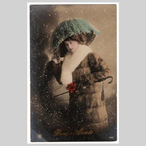Postal vintage con foto real: Mujer, abrigo, sombrero, invierno, moda, glamour, nieve cayendo. Tarjeta de felicitación antigua alemana eduardiana de 1911.
