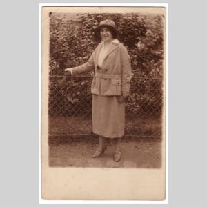 Può includere: Fotografia vintage in tonalità seppia di una persona in un completo e cappello color chiaro, in piedi vicino a una recinzione di rete metallica e fogliame. Il completo presenta una giacca con cintura e tasche e una gonna abbinata. La persona indossa un cappello e scarpe.