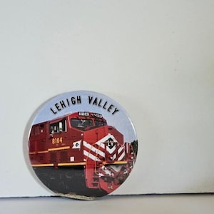 May include: A round button with a red, white, and black train engine with the words "Lehigh Valley" printed in black on a white background.