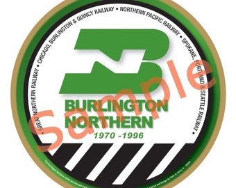 May include: A round, gold-rimmed ornament featuring a green and white logo for the Burlington Northern Railway. The logo displays the letters "BN" in a stylised font. The text "Burlington Northern" and "1970-1996" are printed beneath the logo. The ornament is decorated with black and white stripes.