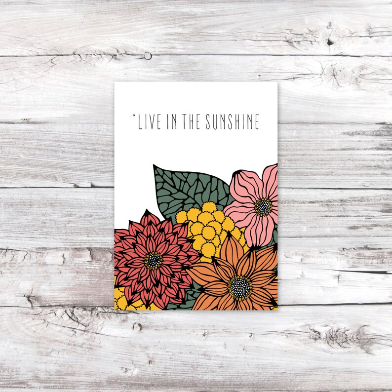 May include: A white card with the text "LIVE IN THE SUNSHINE" printed in black. The card is decorated with a cluster of colorful flowers, including pink, orange, yellow, and red.
