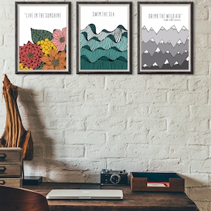 May include: Three framed prints with inspirational quotes and illustrations. The first print features a floral design with the quote "Live in the sunshine". The second print features a blue and green wave design with the quote "Swim the sea". The third print features a gray mountain design with the quote "Drink the wild air".