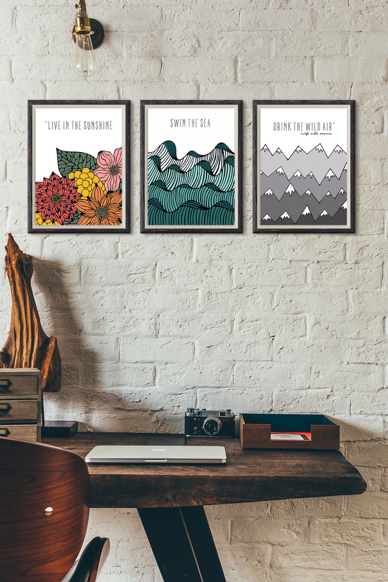 May include: Three framed prints with inspirational quotes and illustrations. The first print features a floral design with the quote "Live in the sunshine". The second print features a blue and green wave design with the quote "Swim the sea". The third print features a gray mountain design with the quote "Drink the wild air".