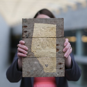 May include: A woman holds a wooden sign with a gold outline of the province of Alberta, Canada. A small heart is painted in gold on the sign.