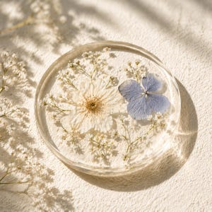 May include: A clear, round resin coaster with dried flowers embedded inside. The coaster features a large white flower, a blue hydrangea petal, and small white flowers. The coaster is on a white surface.