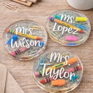May include: Three round, clear resin coasters containing embedded school supplies like miniature pencils, crayons, paper clips, and erasers. Each coaster features white script text reading Mrs. Wilson, Ms. Lopez, and Mrs. Taylor.