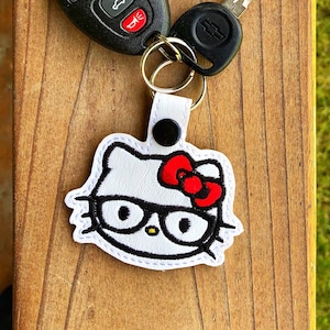 May include: White Hello Kitty keychain with black glasses and a red bow. The keychain is attached to a silver key ring and a black car key with a silver key.