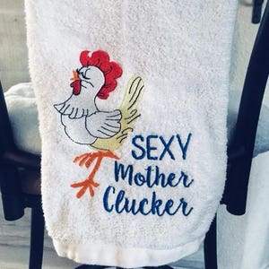 May include: White hand towel with an embroidered cartoon chicken design. The chicken has a red comb, yellow beak and feet, and a white body. The text "SEXY Mother Clucker" is embroidered in blue. The towel has the text "LITTLE SHOP ON THE TUNDRA".