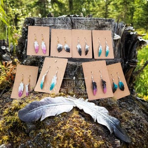 May include: A collection of feather-shaped earrings displayed on brown cardboard cards, featuring various color gradients including pink, white, black, and teal. The text Little Shop on the Tundra is printed in the center. A large grey feather lies below.